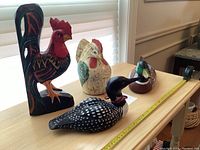 Four bird ornaments including a tall black and red wood rooster, a white and green wood chicken, a mallard duck, and a black and white plastic loon coin bank on a wooden table with measuring tape