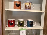 Five ceramic mugs with rooster designs on two white shelf levels, different colored backgrounds and rim patterns visible