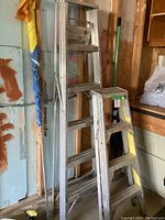 Two aluminum step ladders leaning against a wall. One ladder is approximately 6 feet tall with six steps, the other about 3 feet tall with four steps. Both ladders show use marks and paint splatters.