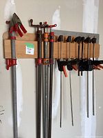 Photo showing 6 bar clamps and 4 pipe clamps hanging on a wooden rack in a garage, displaying the assortment and condition of the metal bars, handles, and clamp jaws.