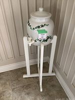 Front view of the white ceramic water dispenser with green ivy design on white wooden stand, showing the spigot and lid.