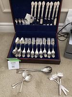Complete flatware set arranged inside a wooden box with dark lining. Includes 8 dinner forks, 8 dinner knives, 8 soup spoons, 3 dessert forks, 3 dessert spoons, salad tongs, and additional small spoons laid outside the box.