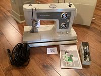 Kenmore sewing machine with power cord, instruction manual, and box of accessories on wooden floor.