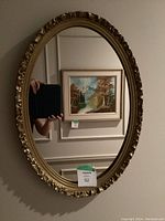 Oval mirror hanging on a wall reflecting a painting of a natural landscape with trees and a river. The mirror frame is gold with intricate floral detailing.