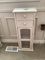 Front view of small white wooden cabinet showing two drawers and glass paneled door.