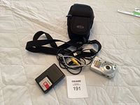 Full overview of the lot items placed on white quilted surface: Sony Cyber-shot camera, charger with battery inserted, carrying case, USB and AV cables, and lot number card.