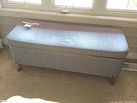 Full view of the light gray upholstered storage bench with padded top, positioned in front of windows.