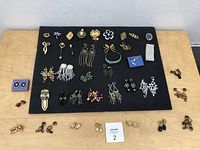 Top-down image showing a black display board filled with assorted vintage costume jewelry including earrings, brooches, and necklace pieces in various shapes and colors.