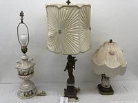 Three vintage Victorian lamps: porcelain base with painted scenes and figurine finial, brass base with porcelain figurine, brass base with figurine finial, all with fabric shades.