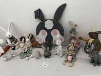 Assorted bunny figurines and plush toys arranged in a semi-circle, including large plush bunnies holding carrots, smaller decorative figurines, and a large black wooden bunny decoration with pink paw pads.