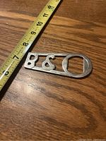 Photo of the Bang & Olufsen bottle opener showing the metal piece with cut-out logo and a measuring tape indicating length on a wooden surface.
