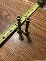Two brass candle holders placed on wooden surface with measuring tape showing height approx 5 and 6 inches respectively.