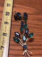 Brooch with blue and green faceted stones next to a measuring tape showing approx 3.5 inch length