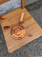 Copper frying pan placed on a wooden chair, showing the entire pan and its handle.