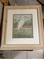 Framed print of white crane bird standing in grass near water with natural wood frame and white mat.