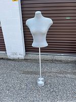 Photo of gray female dressmaker's bust on metal stand with weighted silver round base standing outside on pavement.