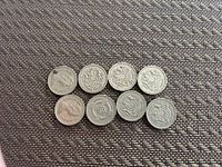 Photo shows 8 English one pound coins laid out in two rows on a textured surface. Various designs visible on the coins including lions and heraldic emblems.