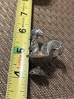 Filigree rooster figurine placed next to measuring tape showing height of about 5.5 cm.