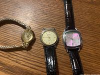 Photo showing three ladies wrist watches on a wooden surface: a gold-tone watch with floral bezel, a round gold-tone watch with black leather strap, and a square silver-tone watch with pink dial and black leather strap.