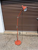 Full view of the mid century floor lamp showing adjustable arm and lampshade in reddish-orange finish, standing on concrete ground in front of a brown garage door.