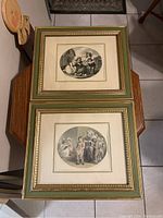 Photo of the pair of framed art print etchings showing the full frames and mats, arranged on a small wooden table.