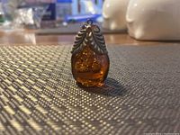 Amber-colored pear-shaped pendant with metal leaf cap photographed on textured surface, showing front detail.