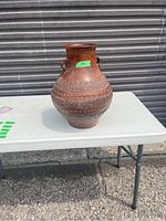 Image of reddish-brown pottery vase with detailed raised designs and two small loop handles, placed on a table outdoors.