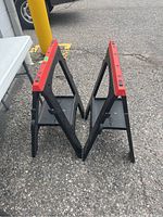 Two black and red plastic saw horses standing side by side on asphalt.