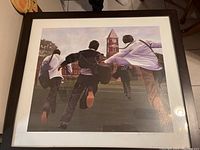 Framed artwork print showing four people running across lawn towards a clock tower building, with dark wooden frame, approximately 25x22 inches.