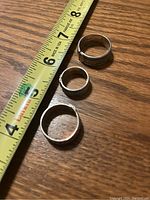 Top down view of three silver rings next to a measuring tape showing sizes approximately 7 to 8 cm in diameter on wooden surface.