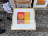 Framed abstract art print displayed on ground with geometric shapes in warm colors and light wood frame.