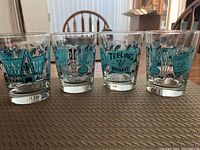 Four short glasses with distinctive blue-green and black tiki-themed prints and Teeling Whiskey branding on a brown textured surface with a wooden chair and partial kitchen background.