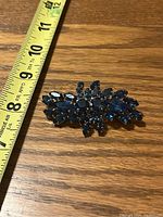 Top-down photo of a women's brooch placed next to a tape measure showing it is approximately 3 inches wide. The brooch features dark blue, faceted rhinestones of various shapes set in silver-tone metal on a wooden surface.