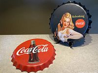 Two Coca-Cola metal bottle cap signs displayed side by side with vintage graphics and logos.