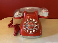 Front view of red Coca-Cola branded rotary style push button phone with snow dome enclosures on handset sides