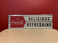 Front view of Coca Cola corrugated tin metal sign with red and white logo and black text on silver corrugated metal.