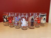 Front view of seven Coca Cola illustrated drinking glasses, showing various designs including polar bears, a dog, paw prints, a bottle, and logos.