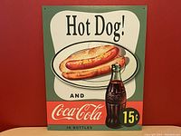 Full front view of Coca Cola Hot Dog tin sign against a red wall.