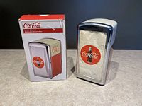 Front view of Coca Cola diner style napkin dispenser next to original box showing branding and product image.