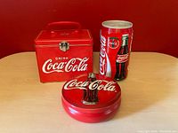 Red Coca Cola tin lunchbox, round tin container, and soda can shaped canister shown on table.