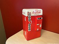 Full view of the Coca Cola vending machine cookie jar standing on a table against a red wall showing front and side vending machine design details.