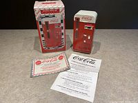 Photo showing the die cast metal Coca-Cola vending machine musical bank, original box with Coca-Cola branding, and instruction papers laid out on a surface.