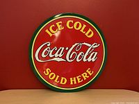 Front view of the round red Coca-Cola embossed metal sign with yellow and green accents placed against a red wall.