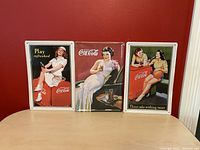 Front view of three Coca Cola tin signs with vintage advertising on a table against red wall