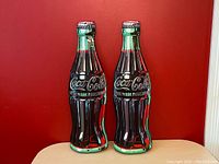 Front view of two Coca-Cola bottle-shaped metal signs, one painted with classic Coca-Cola colors and the other in metallic silver finish.