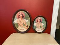 Two oval Coca-Cola trays with woman in pink hat and white dress, placed side by side for scale comparison.