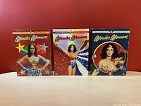 Front view of three Wonder Woman DVD box sets for seasons 1-3 against a red background.