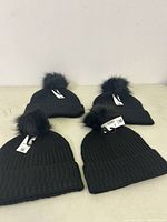 Four black ribbed knit winter hats with fluffy black pom poms, laid on a beige surface.