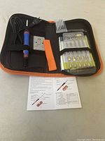 Photo of the wood burning tool kit laid open showing the main pen-style heating tool with blue grip, metal stand, multiple brass tips, orange heat-resistant grip, and instruction manual inside a black and orange carrying case.