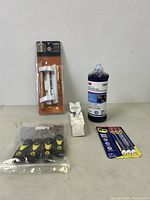 Photo shows pack of 5 small black and yellow scrapers, unopened 3-pack of WD-40 precision pens, 3M grout repair kit bottle, a small white sealed tube, and a white door latch hardware piece on a light surface.